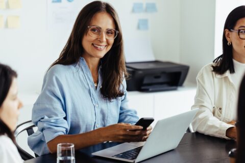 Business woman using a mobile phone during a meeting in an office. Happy business woman sitting in a boardroom with her colleagues.